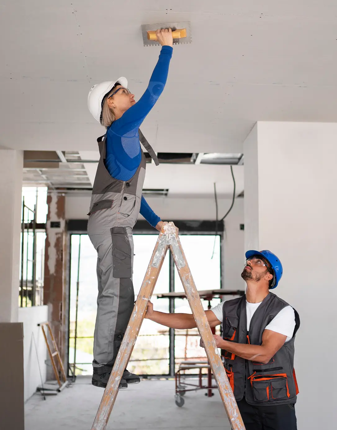 Two construction workers in safety gear work together indoors; one on a ladder applies plaster to the ceiling, while the other supports the ladder, conveying teamwork.