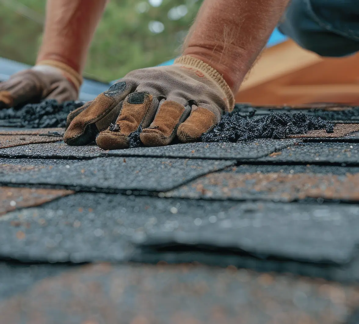 Close-up of hands in work gloves pressing black asphalt shingles onto a roof. The scene conveys focus and effort in roofing construction.