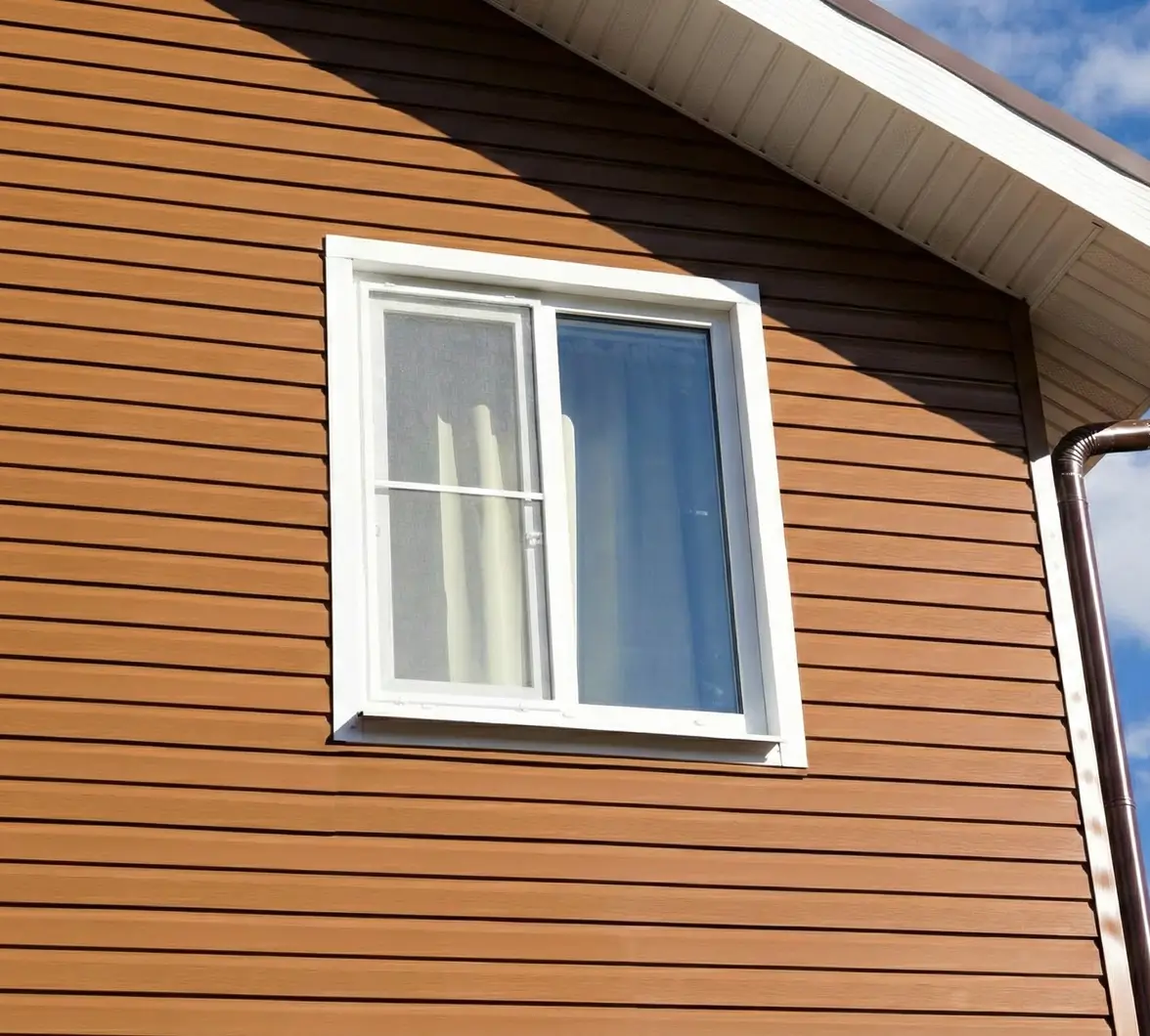 A white-framed window is set in a brown vinyl-sided wall under a clear blue sky. Light curtains are partially visible behind the window glass.