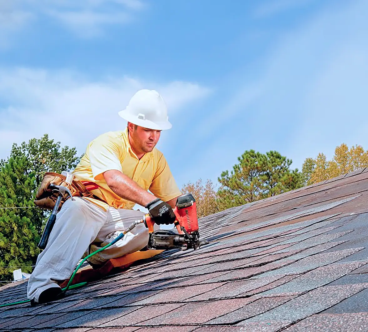 A construction worker in a white hard hat uses a nail gun on a shingled roof, with trees and a blue sky in the background. The scene is bright and clear.