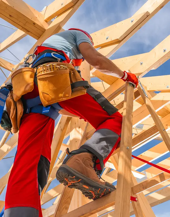 Construction worker in red overalls climbs a wooden roof frame, sky visible above. Tool belt and safety gear emphasize focus on building and safety.