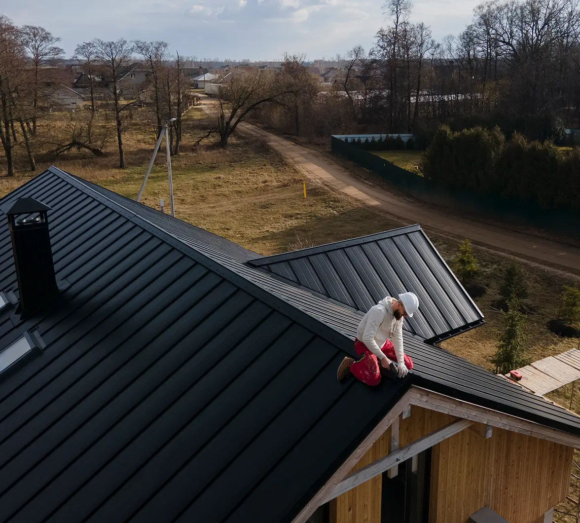 A person in a white jacket and cap works on a dark metal roof of a wooden house, surrounded by bare trees and a dirt road under a cloudy sky.