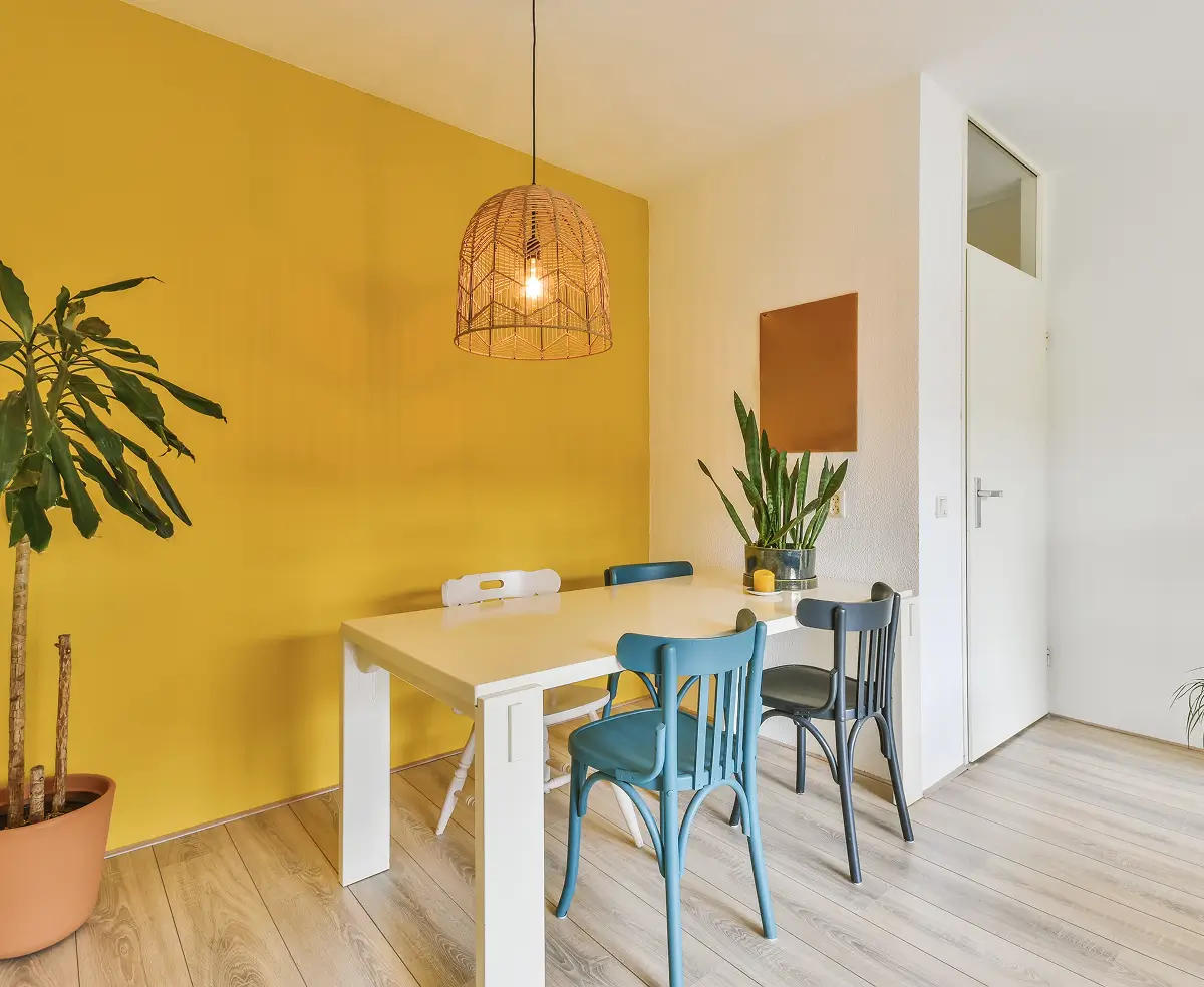 Dining area with a yellow accent wall, a white table, and four mismatched chairs. A woven pendant light, large potted plants, and wooden floor add warmth.