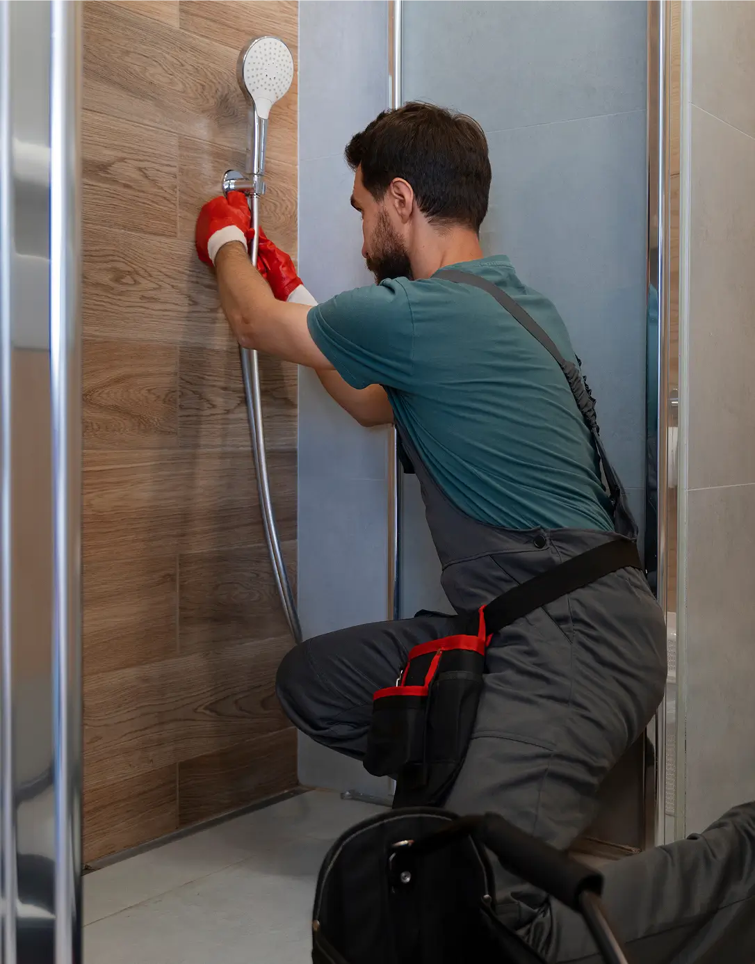 A plumber in overalls kneels in a modern shower, using red gloves to fix a showerhead on a wooden panel wall. A tool bag lies nearby on the floor.