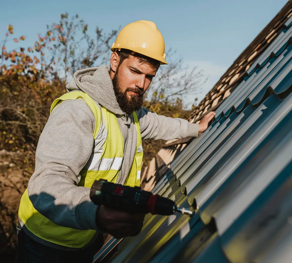 A construction worker in a yellow hard hat and reflective vest uses a power drill to fix metal sheets on a roof. The scene is outdoors on a sunny day.