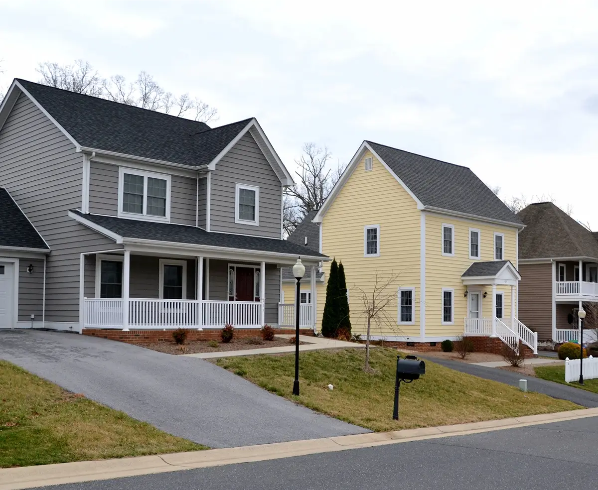 Suburban neighborhood with three houses. A gray house with a porch, a yellow house, and another partially visible house. Overcast sky and tidy lawns convey a calm atmosphere.