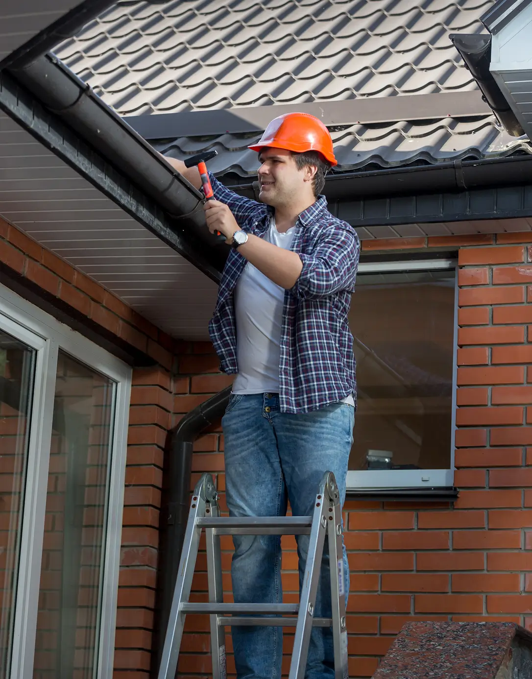 A man in a hard hat stands on a ladder inspecting a house gutter. He wears a plaid shirt and jeans, with a focused expression against a brick wall backdrop.