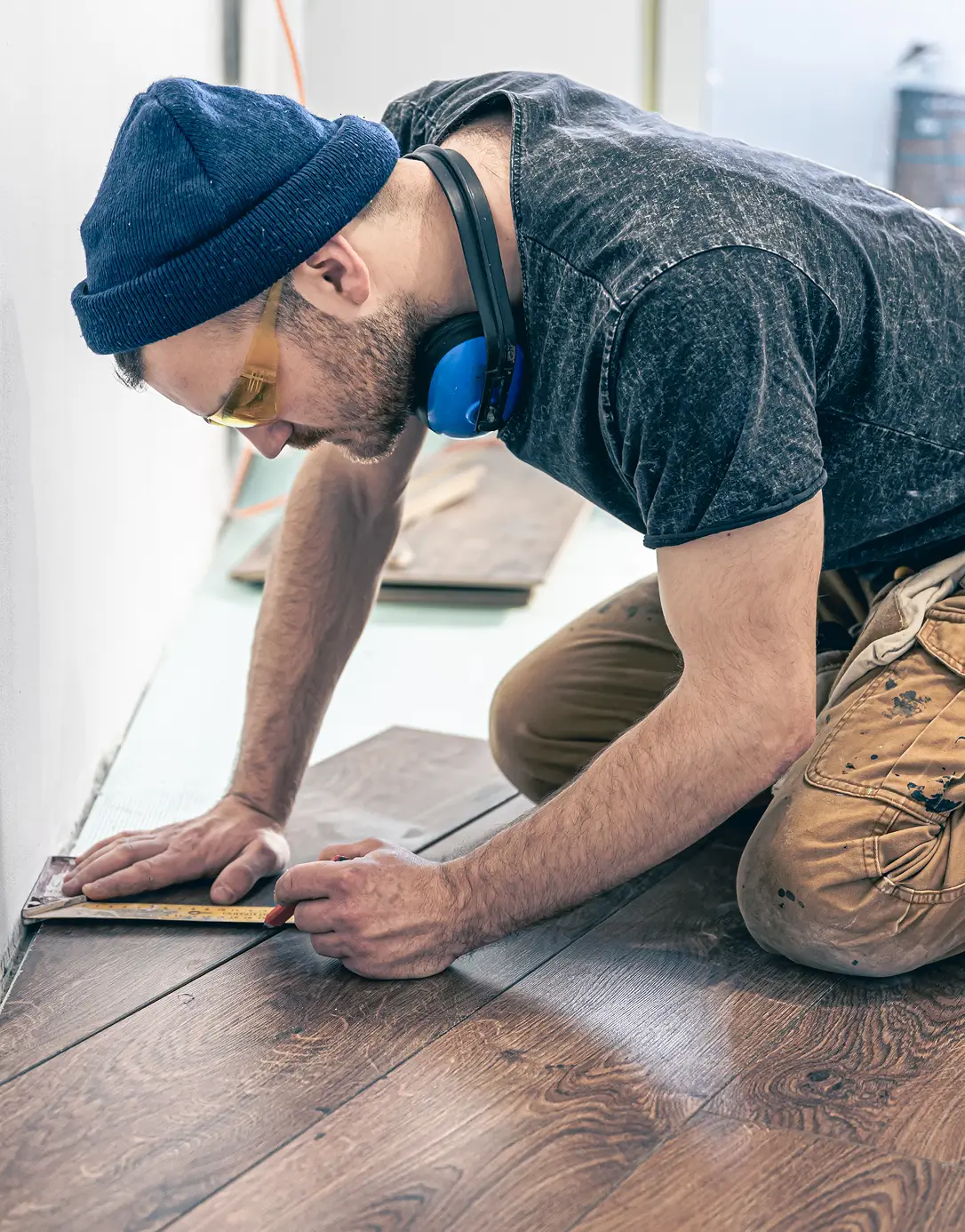 A man wearing a beanie, headphones, and goggles is kneeling, carefully measuring and installing hardwood flooring. His focused expression conveys precision and concentration.