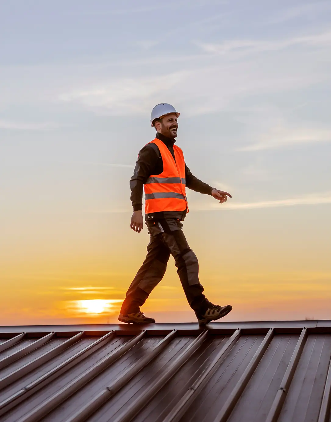 A person in a hard hat and orange safety vest confidently walks on a metal roof at sunset, with a warm sky suggesting a sense of accomplishment.