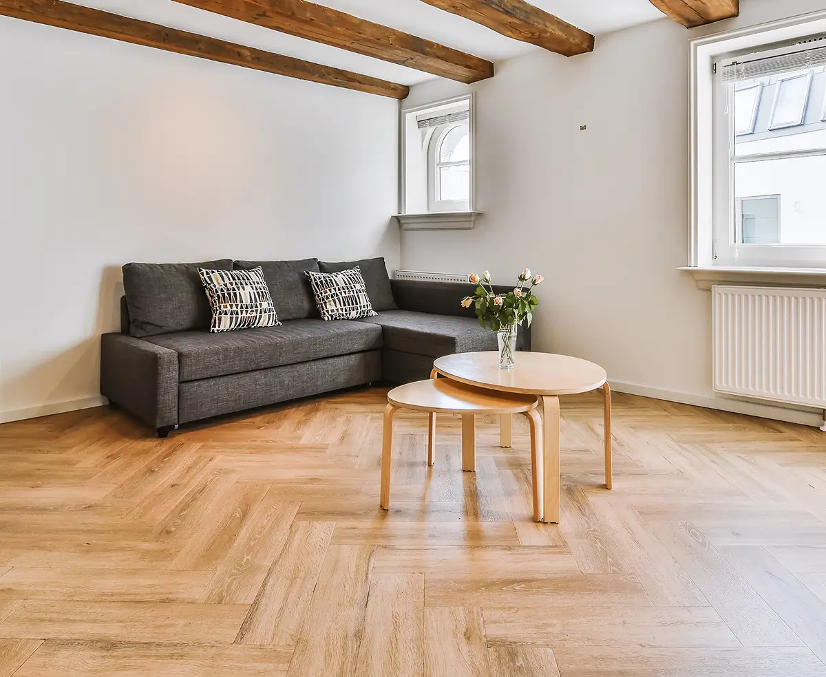 A minimalist living room with a gray L-shaped sofa and patterned cushions. Wooden beams and herringbone floor add warmth. Nesting tables hold flowers.