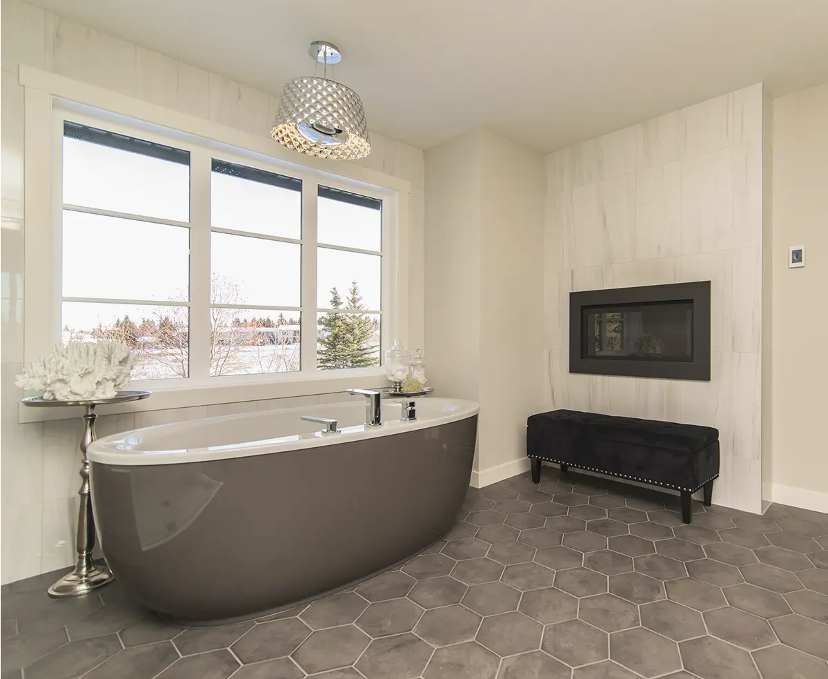 A modern bathroom with a sleek gray bathtub, hexagonal tiles, wall-mounted fireplace, large window with a snowy view, and elegant pendant light.