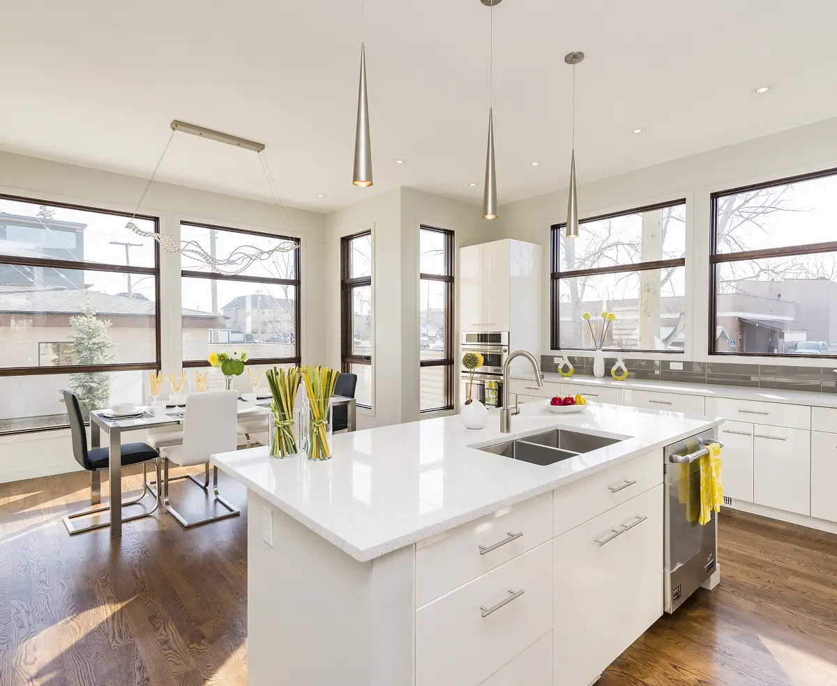 Modern kitchen with white cabinets, sleek island, and pendant lights. Large windows brighten the space, while yellow accents add a cheerful touch.