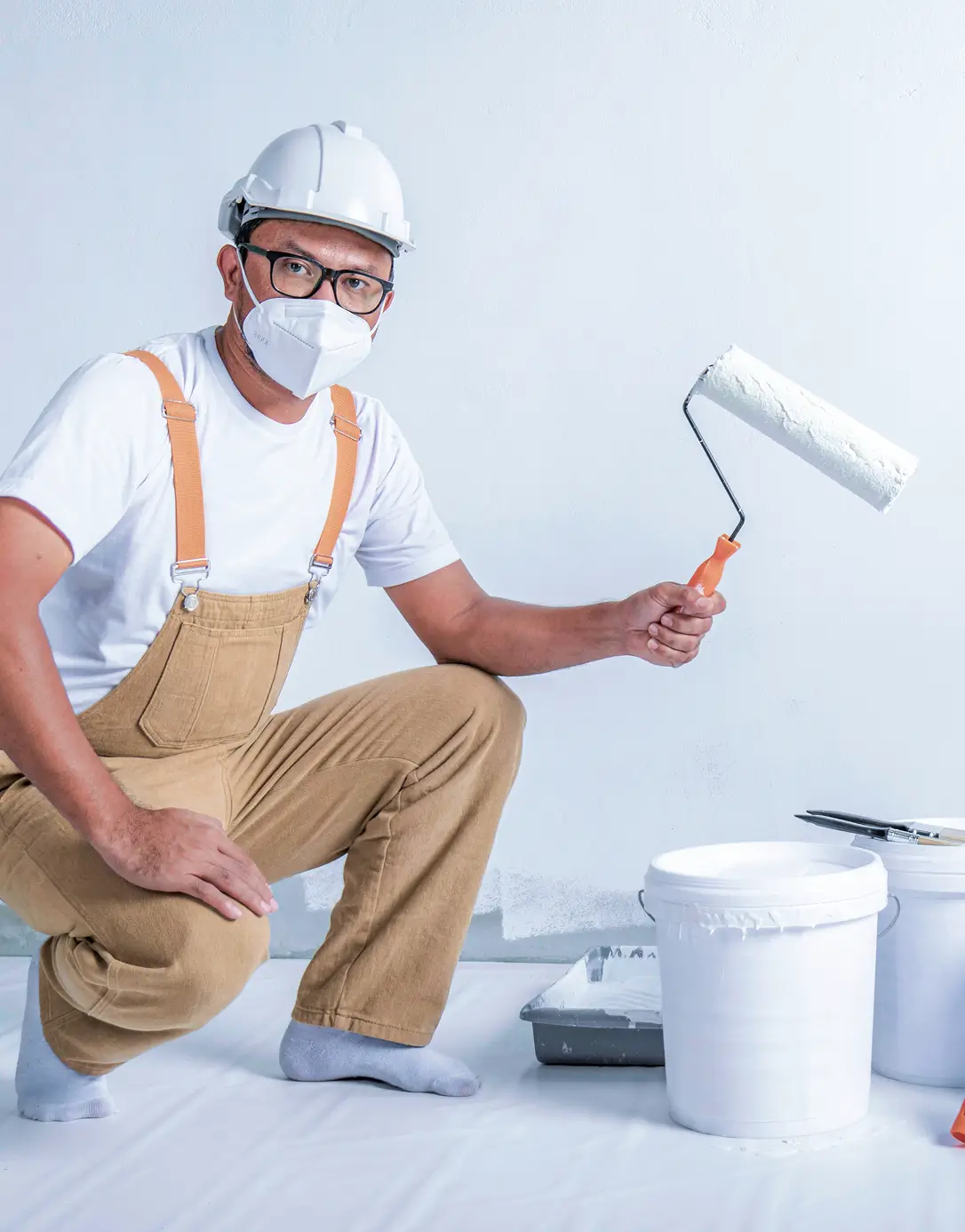 A painter wearing glasses, a white hard hat, and a face mask crouches, holding a paint roller. He wears a white shirt and tan overalls, with paint buckets and tray nearby.