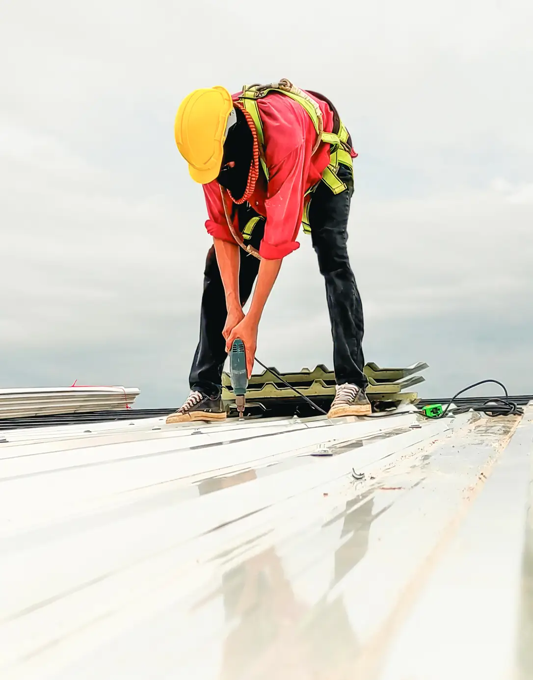 A construction worker in a yellow hard hat and red shirt uses a power drill on a metal roof. The sky is overcast, creating a focused, industrious mood.