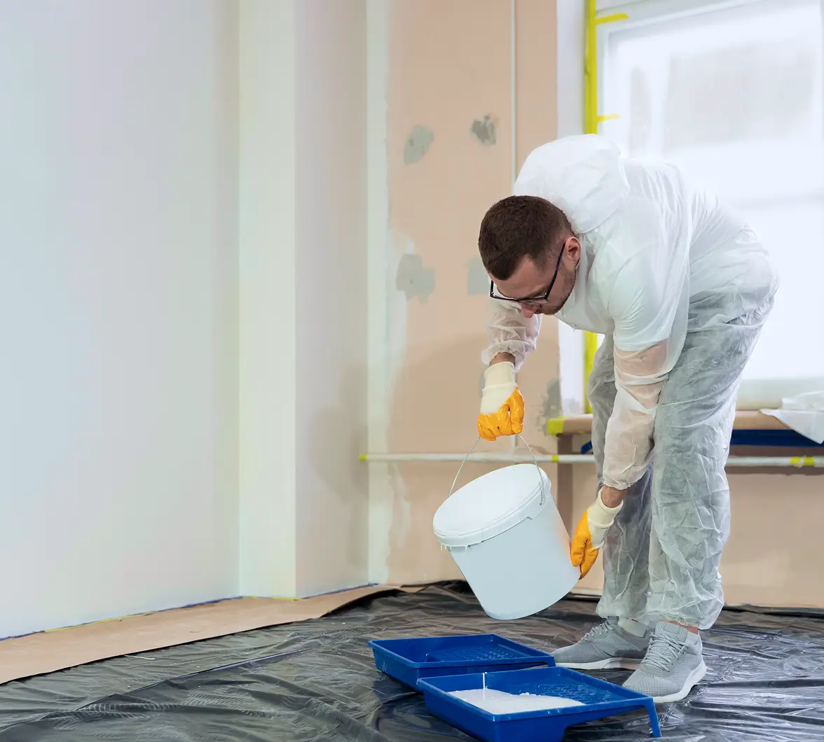 A man in protective gear and gloves pours white paint from a bucket into a tray. Plastic sheets cover the floor, suggesting a room painting in progress.