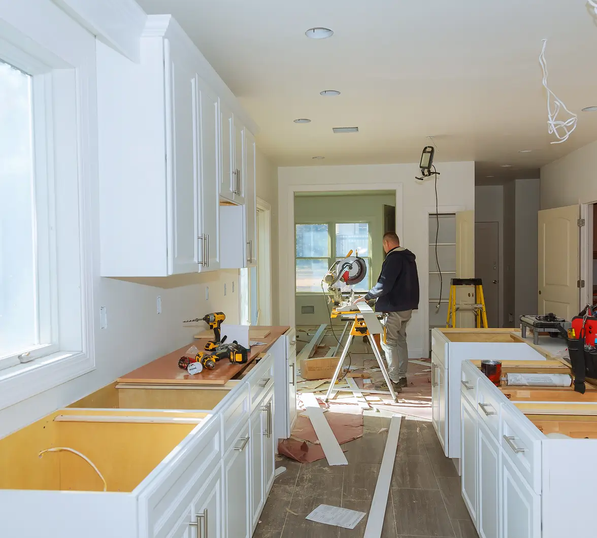 A person working with tools in a bright, under-renovation kitchen with white cabinets. Wood planks and a saw are visible, conveying a focused, busy atmosphere.