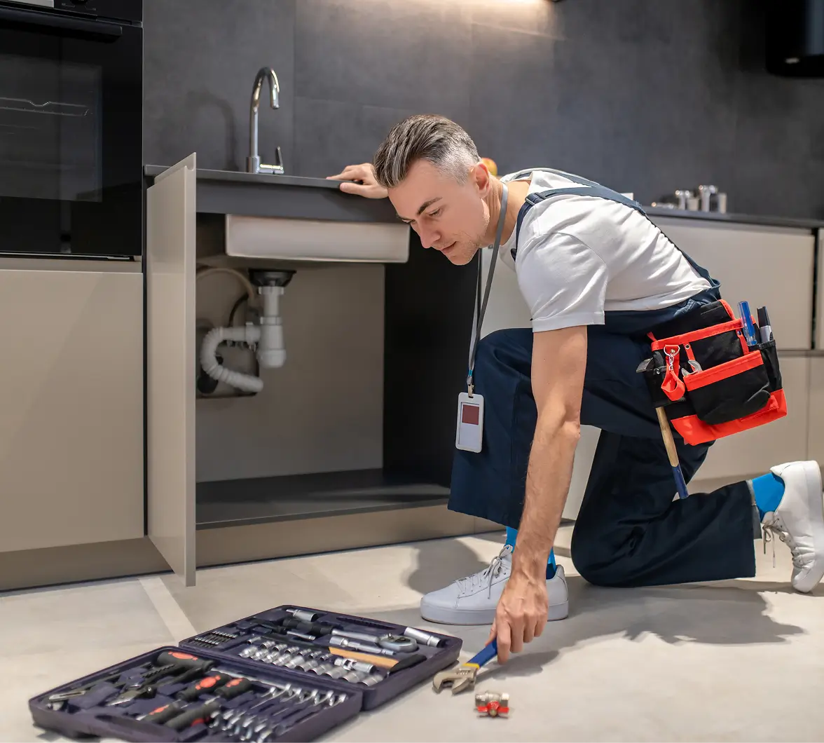 Repairman kneels on a kitchen floor, selecting a tool from an open toolkit. He wears a tool belt and inspects pipes under the sink. Concentrated mood.