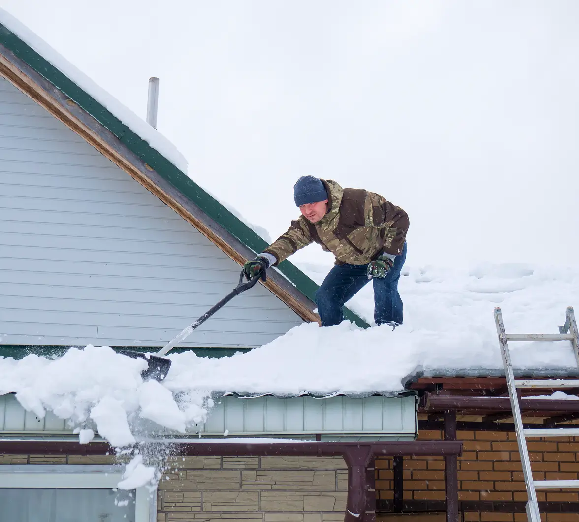 Man in winter clothing shovels snow off a sloped roof, standing near a ladder. Overcast sky; conveys chilly, determined atmosphere.