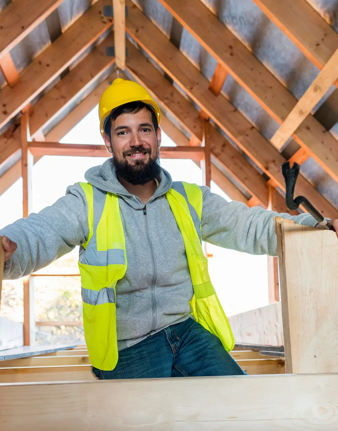 Smiling construction worker in a yellow vest and hard hat sits inside a wooden house frame, conveying a positive and industrious atmosphere.