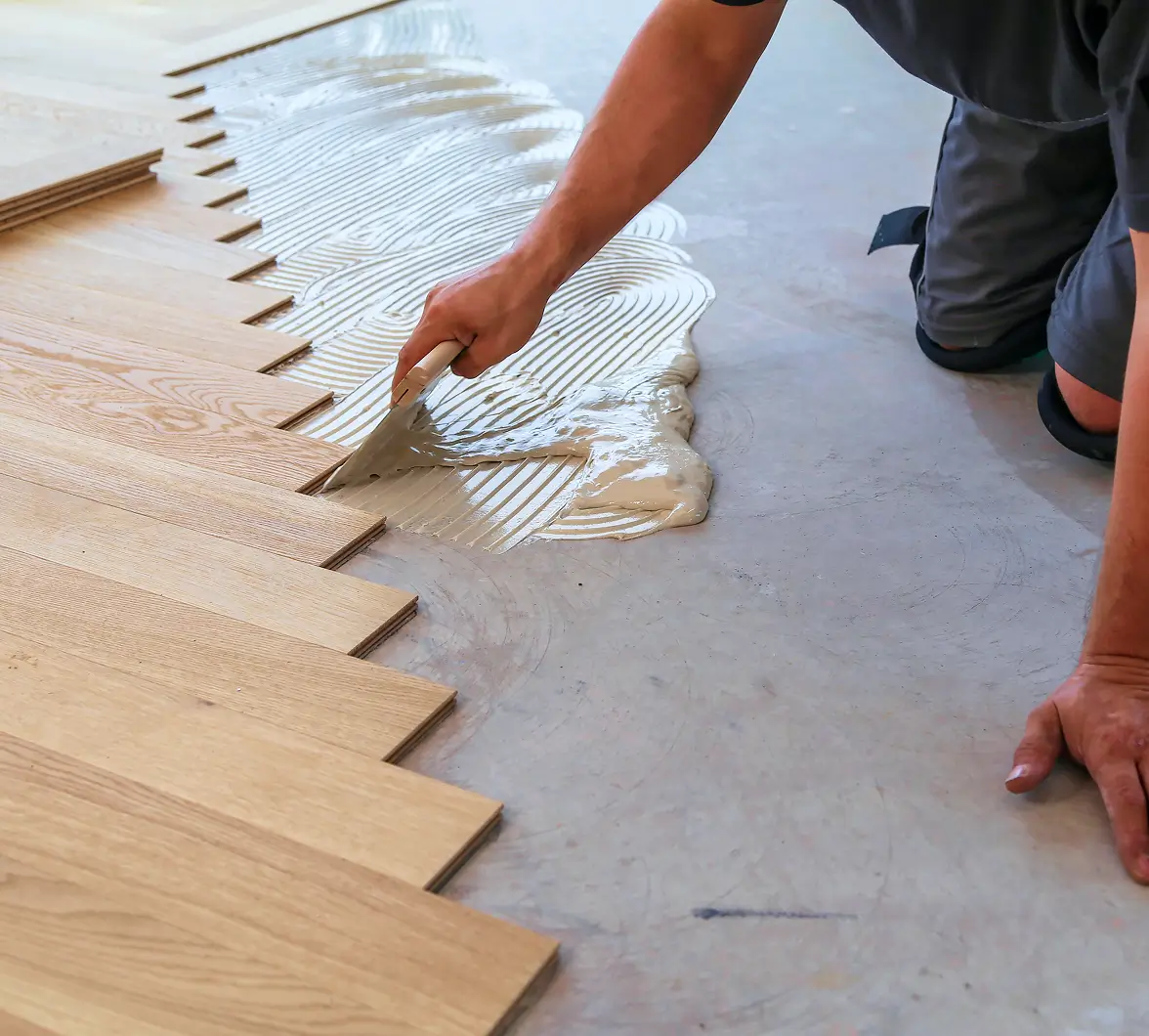 A person is kneeling on the floor, spreading adhesive with a trowel for installing light wooden floorboards. The image conveys precision and craftsmanship.
