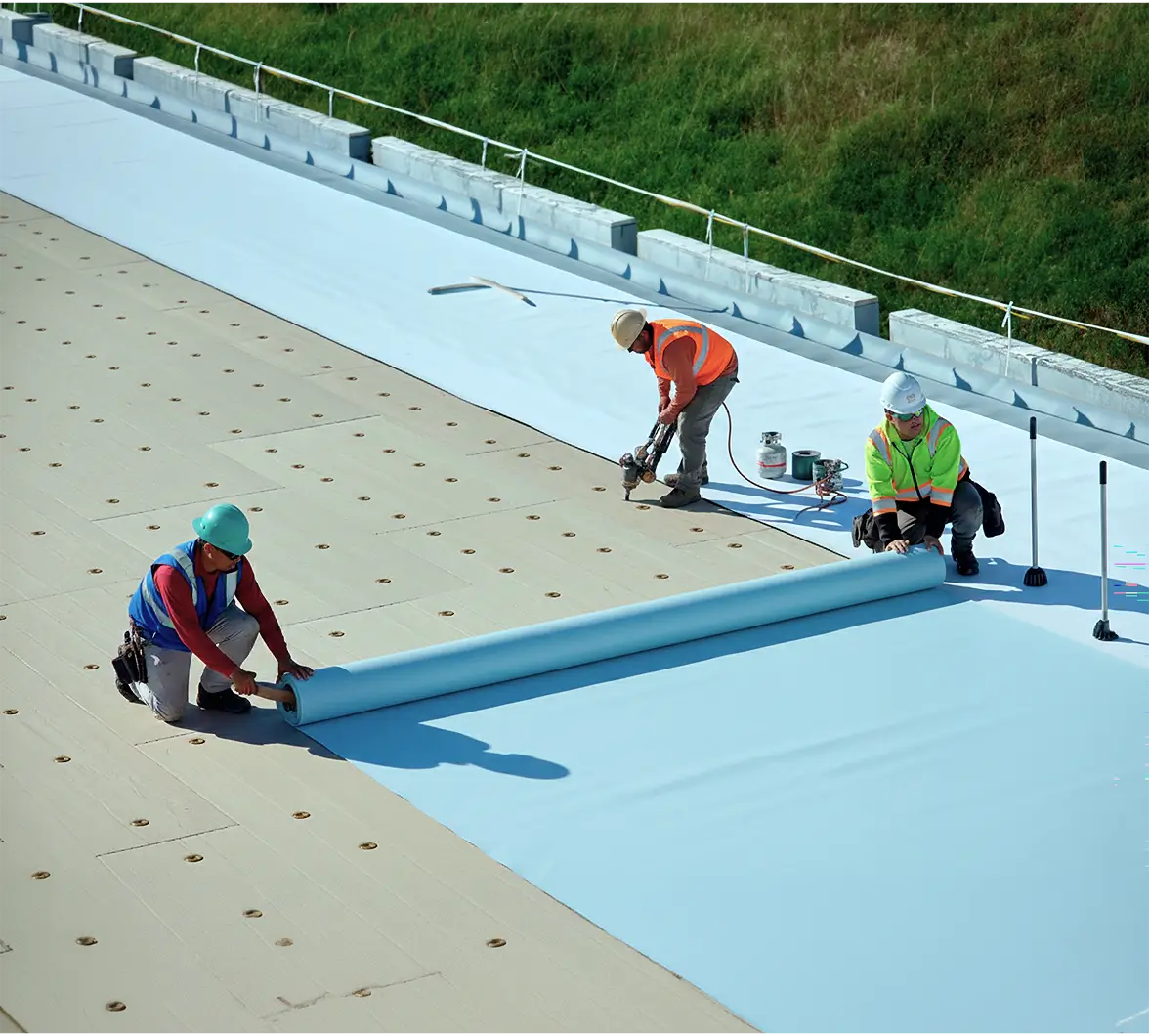 Three construction workers in hard hats and vests lay down blue roofing material on a flat roof, focusing intently on their task under clear skies.
